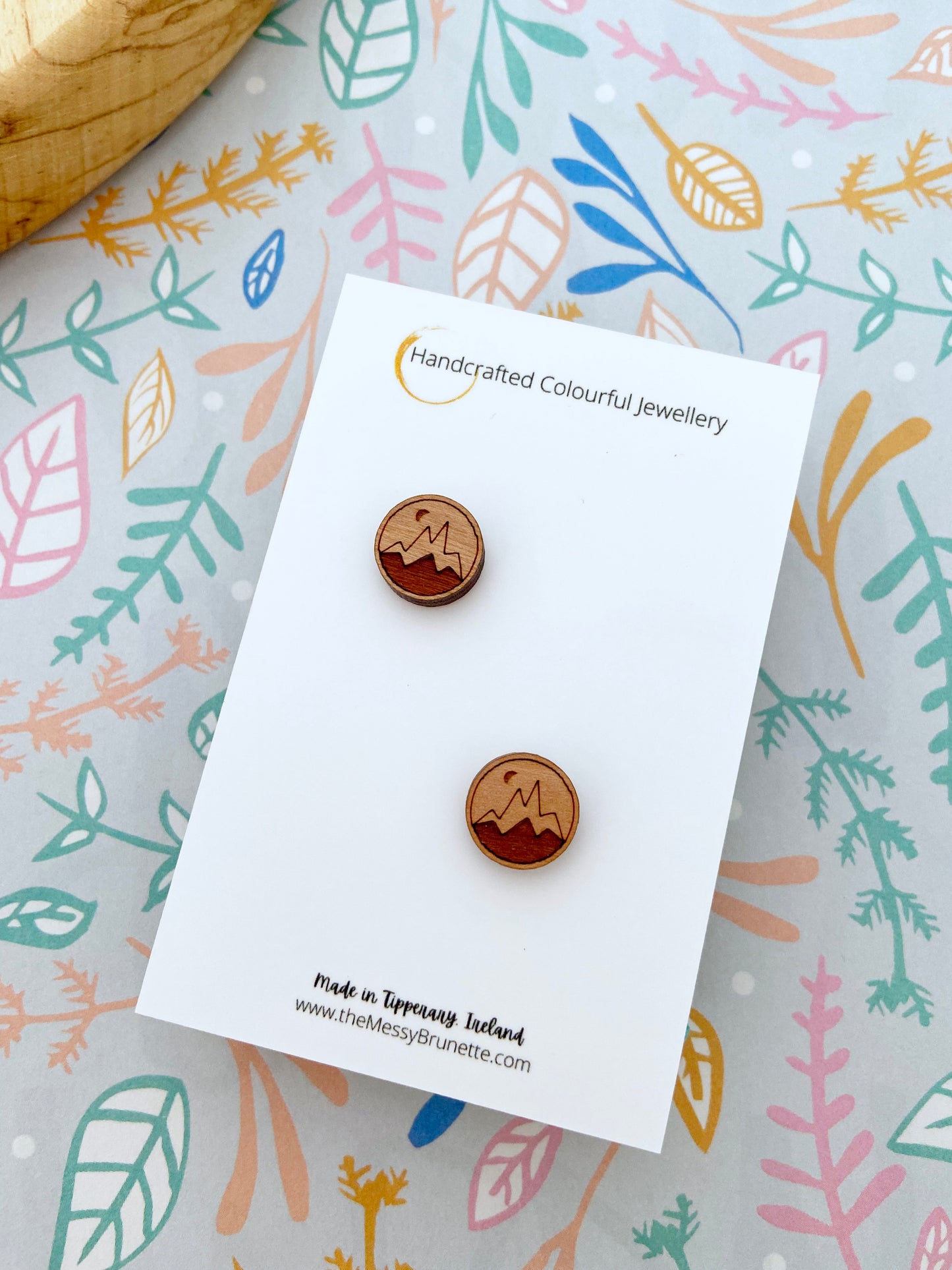 Small wooden stud earrings with a mountain design on them on a branded jewellery card with a colourful leaf pattern in the background