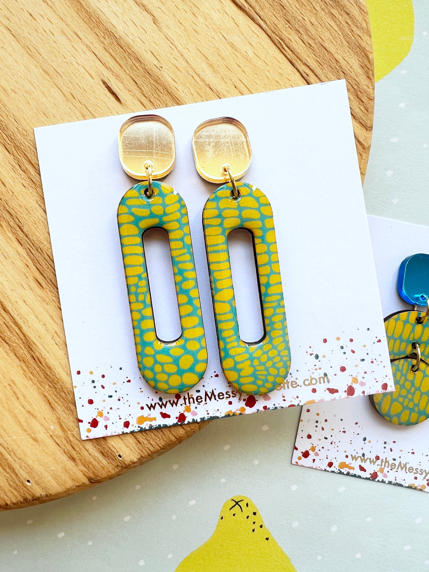 pair of long Yellow and blue patterned earrings on a branded card with a wooden surface background