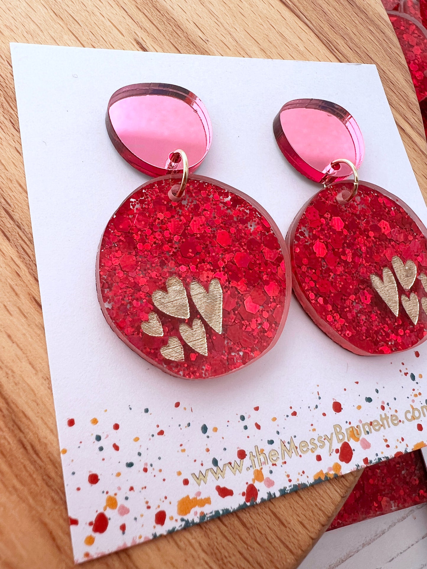 Pink glittery heart earrings on a branded card with a wooden surface background