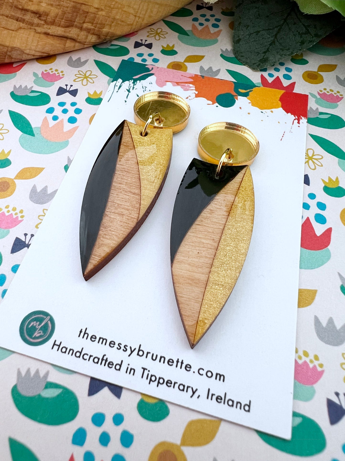 Pair of gold and black painted wood earrings on a colourful patterned card with 'themessybrunette.com' branding.