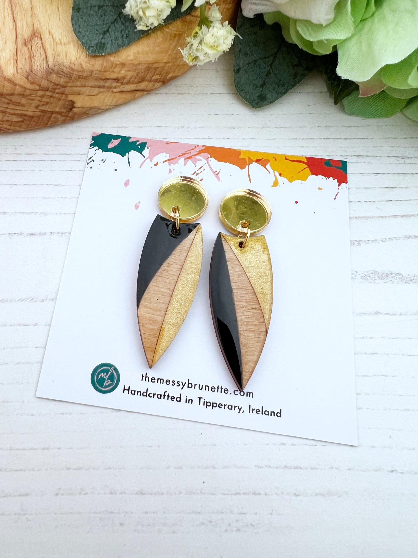 Pair of gold and black wood earrings on a branded card featuring the messy brunette with flowers in the background.
