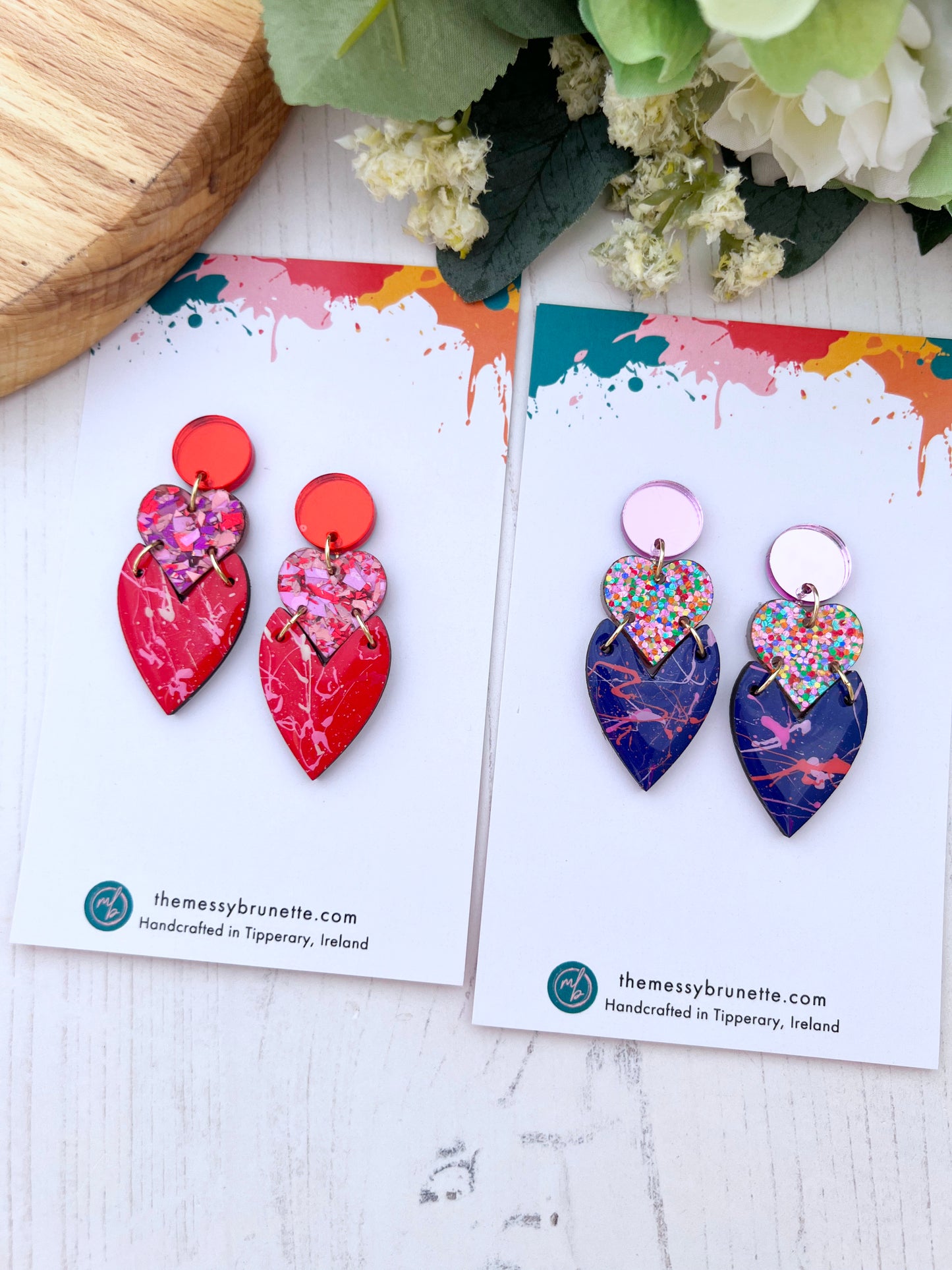 Two pairs of colourful heart-shaped earrings on branded The Messy Brunette cards with a floral background.