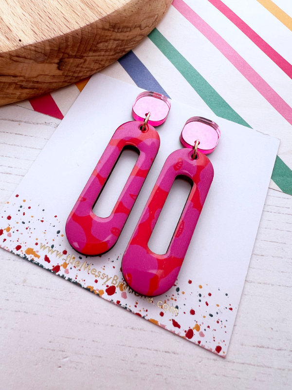 Pair of pink and red patterned earrings on a colorful card with a wooden surface.