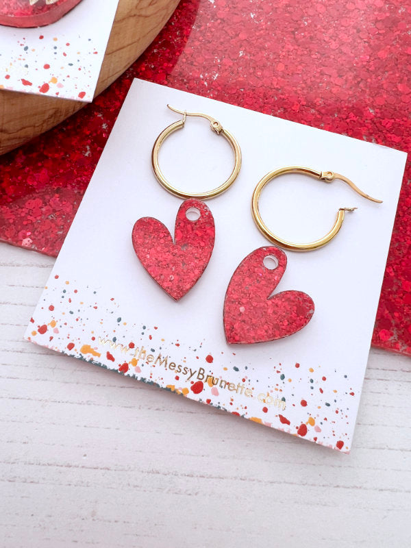 Red heart-shaped earrings on a branded card with a red glittery background
