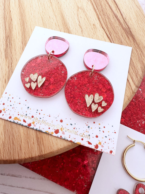 Red glittery earrings with heart designs on a white card with colorful speckles, placed on a wooden surface.