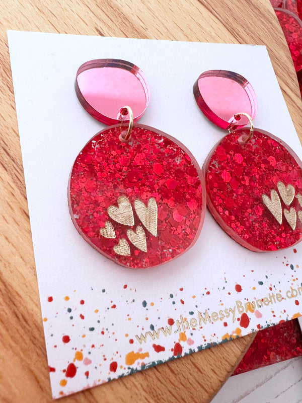 Red glittery heart earrings on a branded card with a wooden surface background
