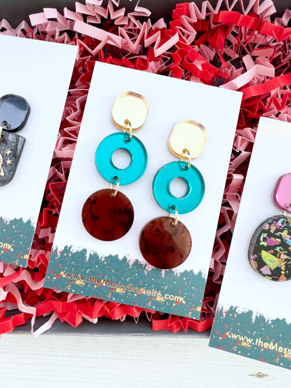 Colorful earrings on a card with 'The Messy Craft Lab' branding, placed on red and pink textured paper.