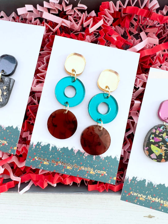 Colorful earrings on a card with 'The Messy Craft Lab' branding, placed on red and pink textured paper.