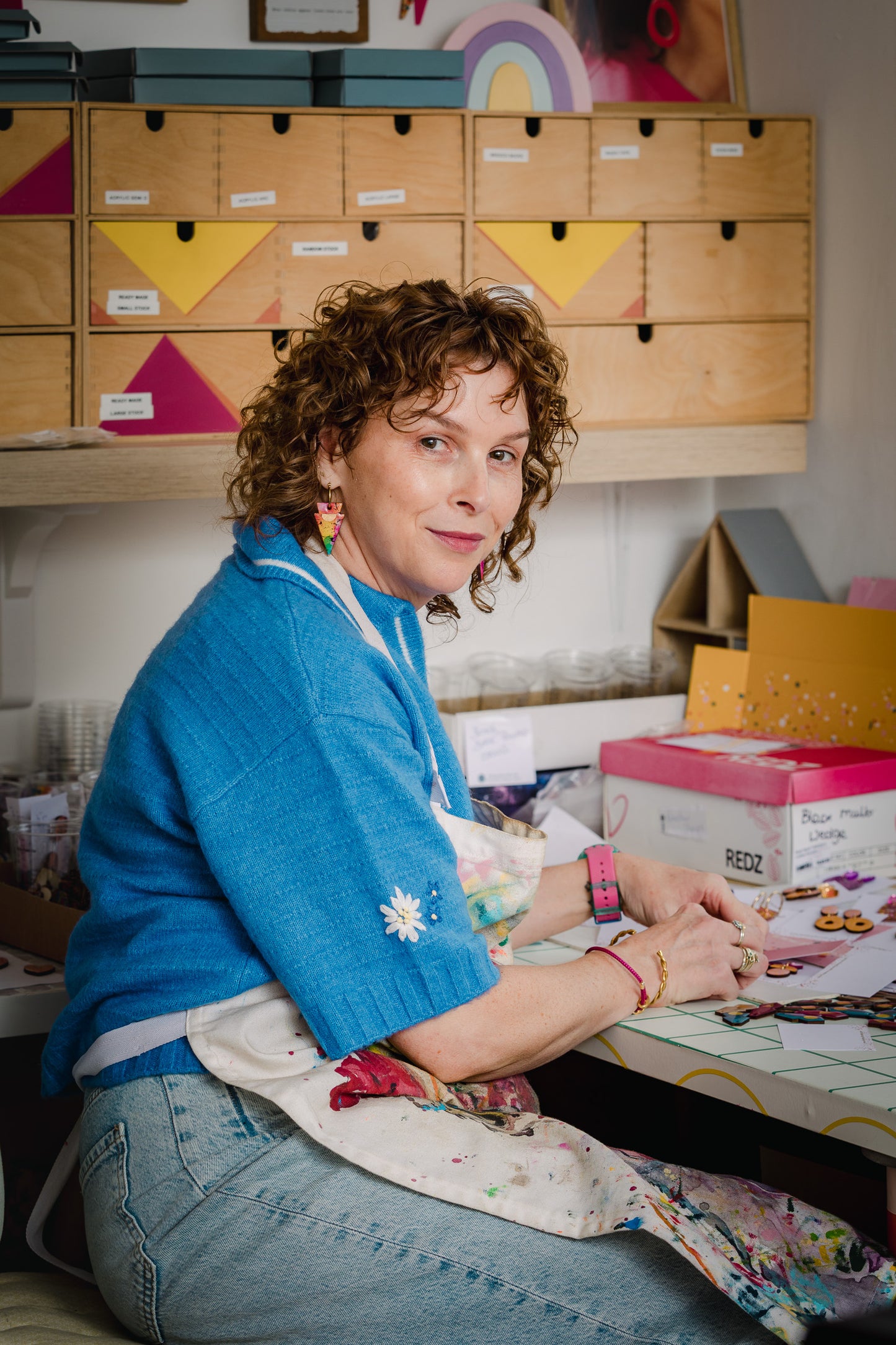 A woman with curly brown hair sits at a table in a colourful craft room, wearing a bright blue cardigan and a white apron with paint splatters. She is surrounded by wooden storage drawers, boxes, and art supplies.