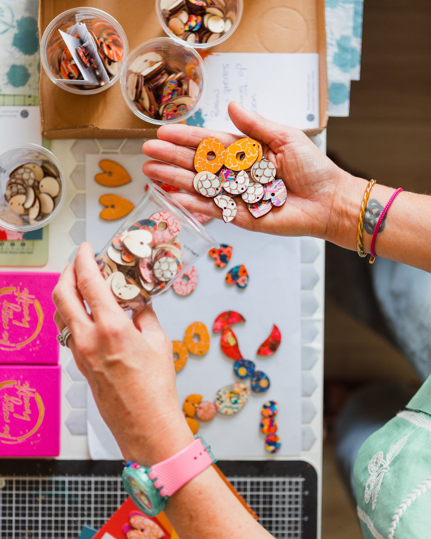 A close-up of hands holding and sorting colourful decorative wood earring pieces, including donut-shaped embellishments and small wooden charms, over a white work surface with more materials and containers.
