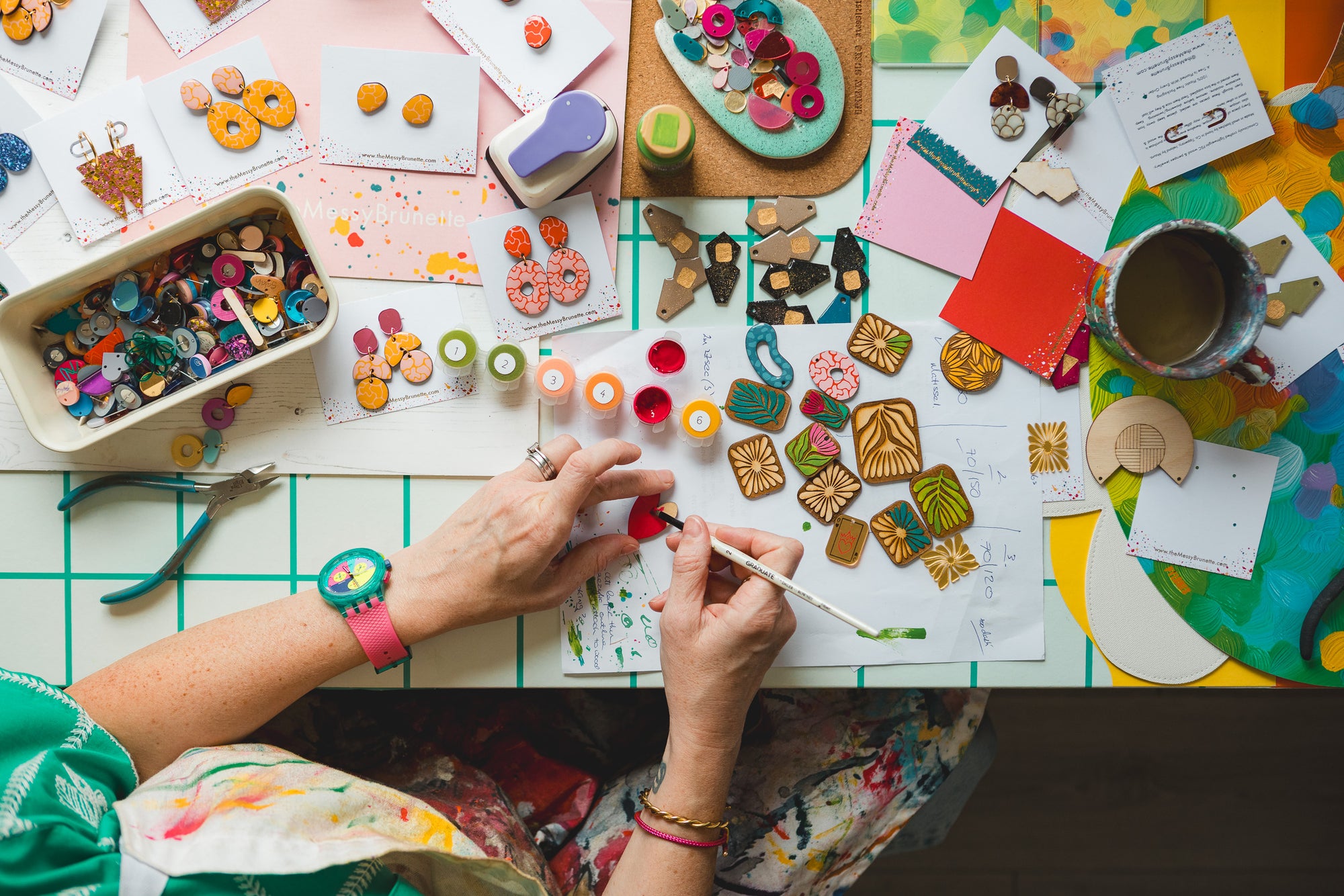 Overhead view of a person's hands painting on a white canvas at a coluorful craft table filled with earrings, paints, stickers, papers, and art supplies.