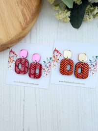Two pairs of geometric drop earrings displayed on white cards on a white wood surface. The left pair features pink and red patterned acrylic drops, while the right pair features gold and orange patterned acrylic drops.